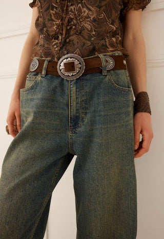 a-close-up-of-a-person-wearing-blue-denim-jeans-with-a-brown-leather-belt-featuring-a-large-silver-toned-western-style-concho-buckle-paired-with-a-patterned-brown-top