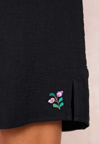 close-up-of-a-black-textured-garment-likely-a-dress-or-tunic-featuring-a-small-embroidered-purple-flower-and-green-leaves-near-the-hem-worn-by-a-model