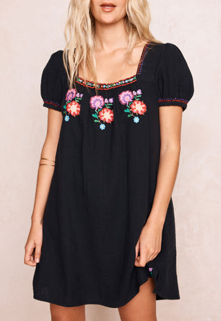 a-woman-wearing-a-black-short-sleeve-dress-with-colorful-floral-embroidery-around-the-neckline-and-on-the-sleeves-photographed-against-a-light-beige-textured-background